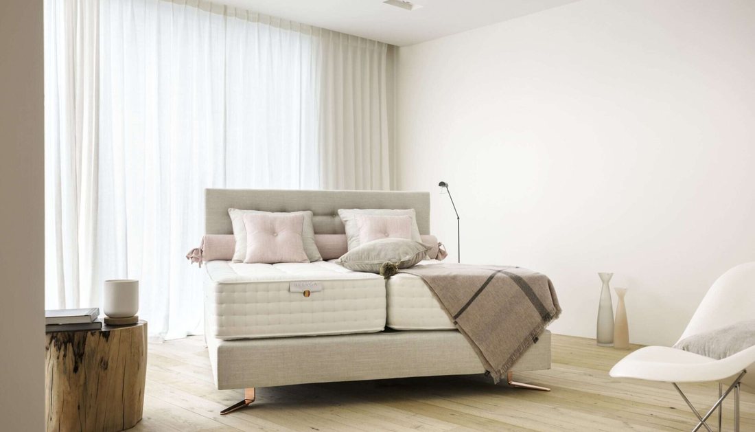 Boxspringbett in einem hellen Raum: gepolstertes Kopfteil, zwei getrennte Matratzenhälften auf dem Unterbau, Tageslicht durch einen Vorhang.