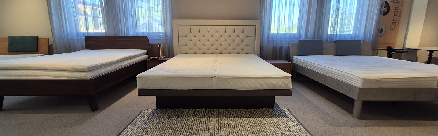 Drei Betten im Showroom nebeneinander, aus derselben Blickrichtung fotografiert; vor dem mittleren Bett liegt ein Handwebeteppich.