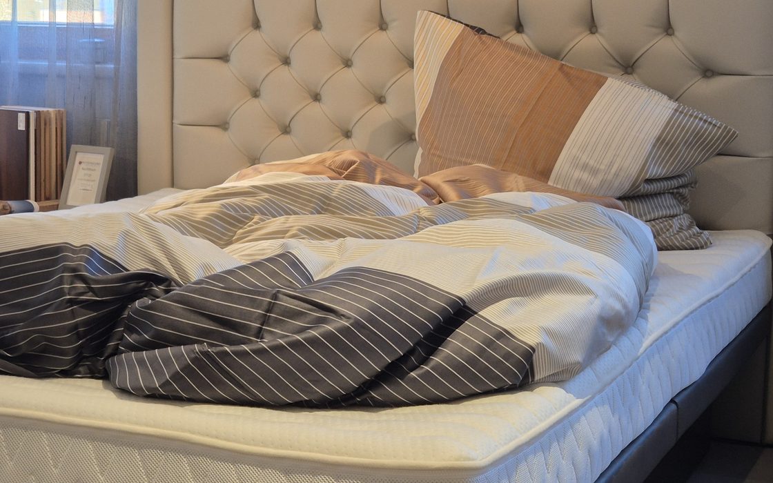 Bezogenes Bett im Showroom: gepolstertes Kopfteil, Kissen und eine locker zurückgeschlagene Zudecke