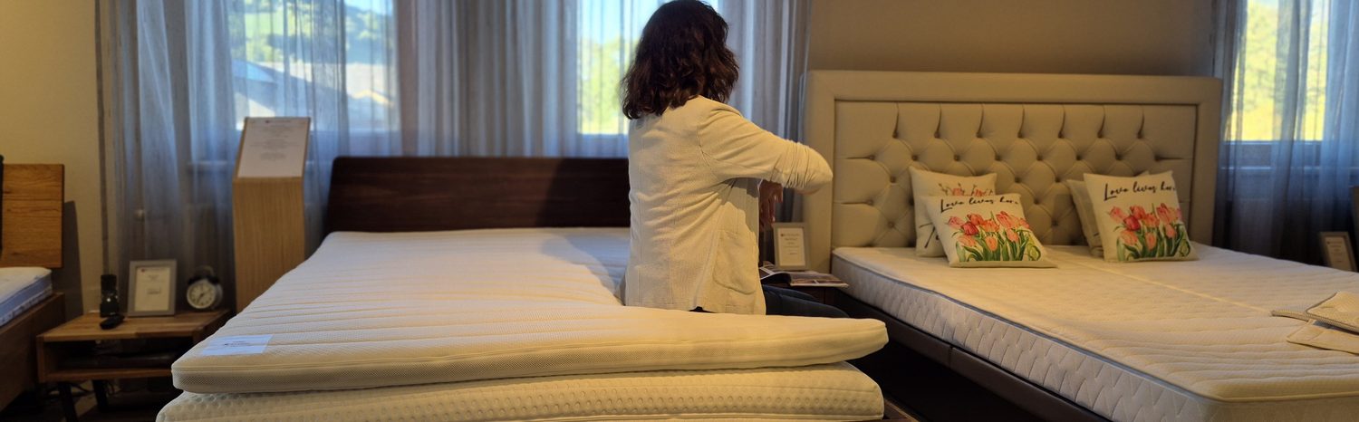 Eine Person sitzt am Rand eines Bettes und schaut zum Fenster hinaus, Morgenlicht im Raum.