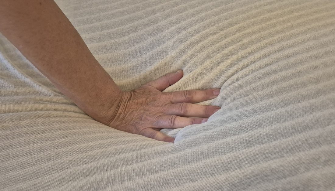 Eine Hand drückt in eine Kaltschaummatratze; der Schaum gibt sichtbar nach, ringsum verläuft der wellenförmige Schnitt der Oberfläche.