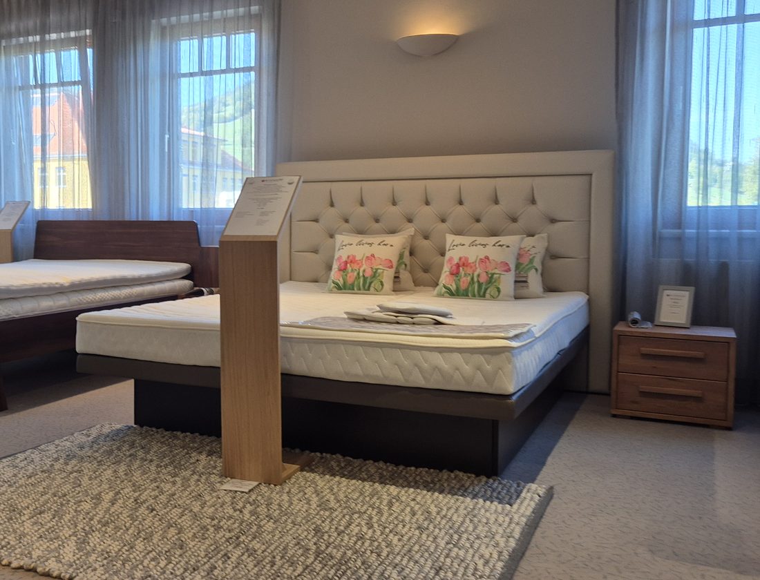 Bett mit gepolstertem Kopfteil und Nachtkästchen im Obergeschoß des Showrooms, davor ein Handwebeteppich